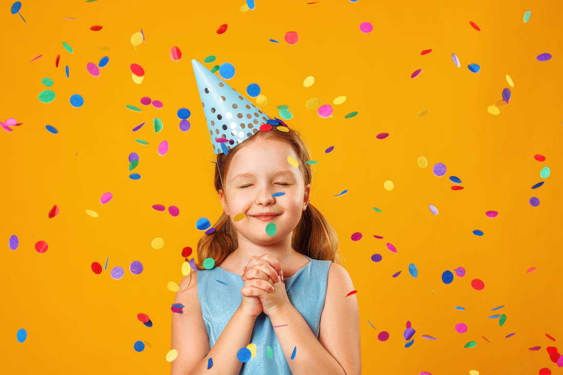 Cute little girl celebrates birthday. The child closed his eyes and makes a wish in the rain of confetti. Closeup portrait on yellow background.