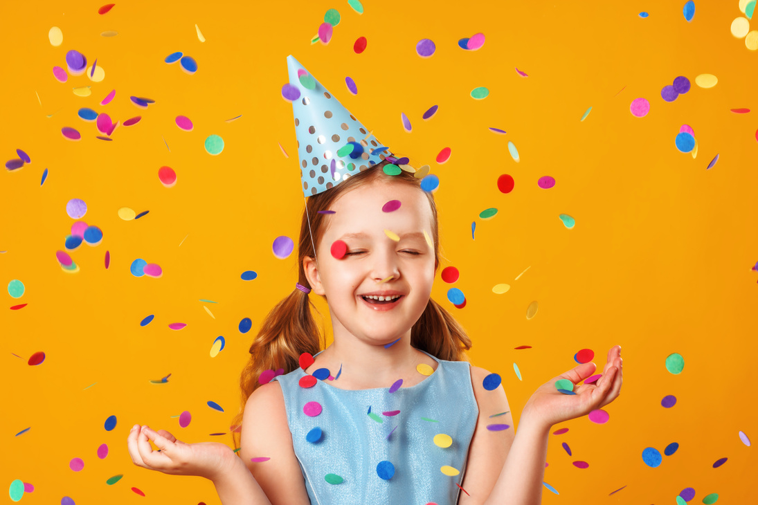 Cheerful little girl celebrates birthday. The child is standing with eyes closed in the rain of confetti. Closeup portrait on yellow background.